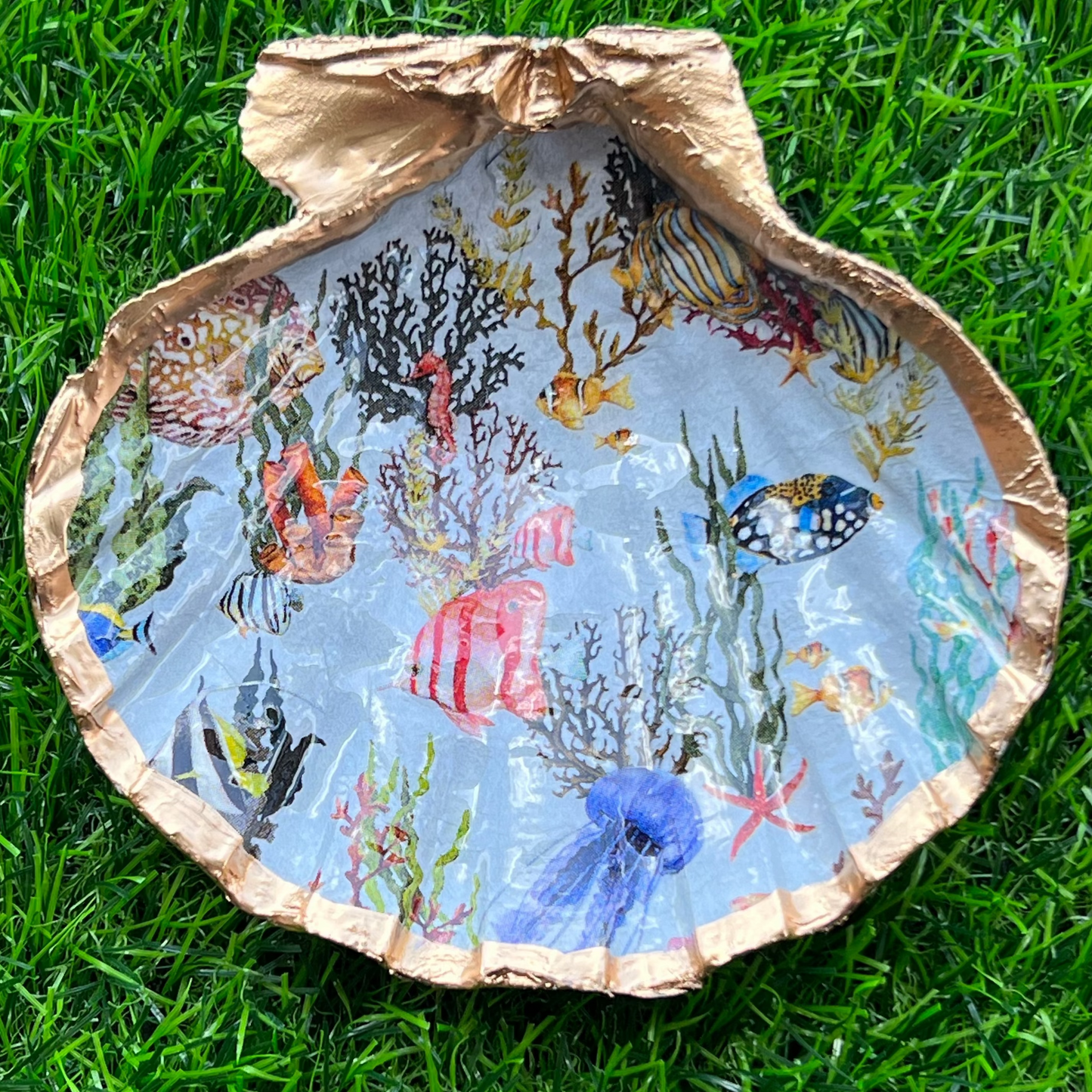 A decorative tray with a blue background featuring a shell design with various colorful sea creatures such as fish and coral, sealed in resin and trimmed in gold leaf, placed on a grass-like surface.