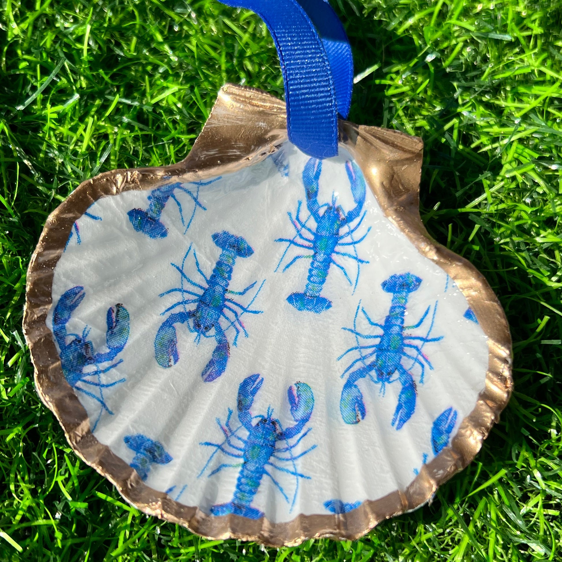 A decorative blue lobster shell with a white background, featuring a pattern of tiny blue lobsters, placed on a grassy surface.
